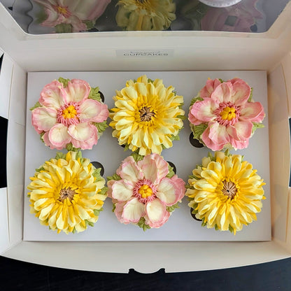 Six handpiped  flower cupcakes arranged in a white square box, featuring pink and yellow dahlia-style blooms with green centers and leafy tops.