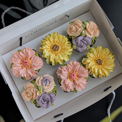A white gift box containing six decorative hand piped floral cupcakes in pastel pink, yellow, peach, and lavender, arranged on a dark surface with a blurred floral foreground.