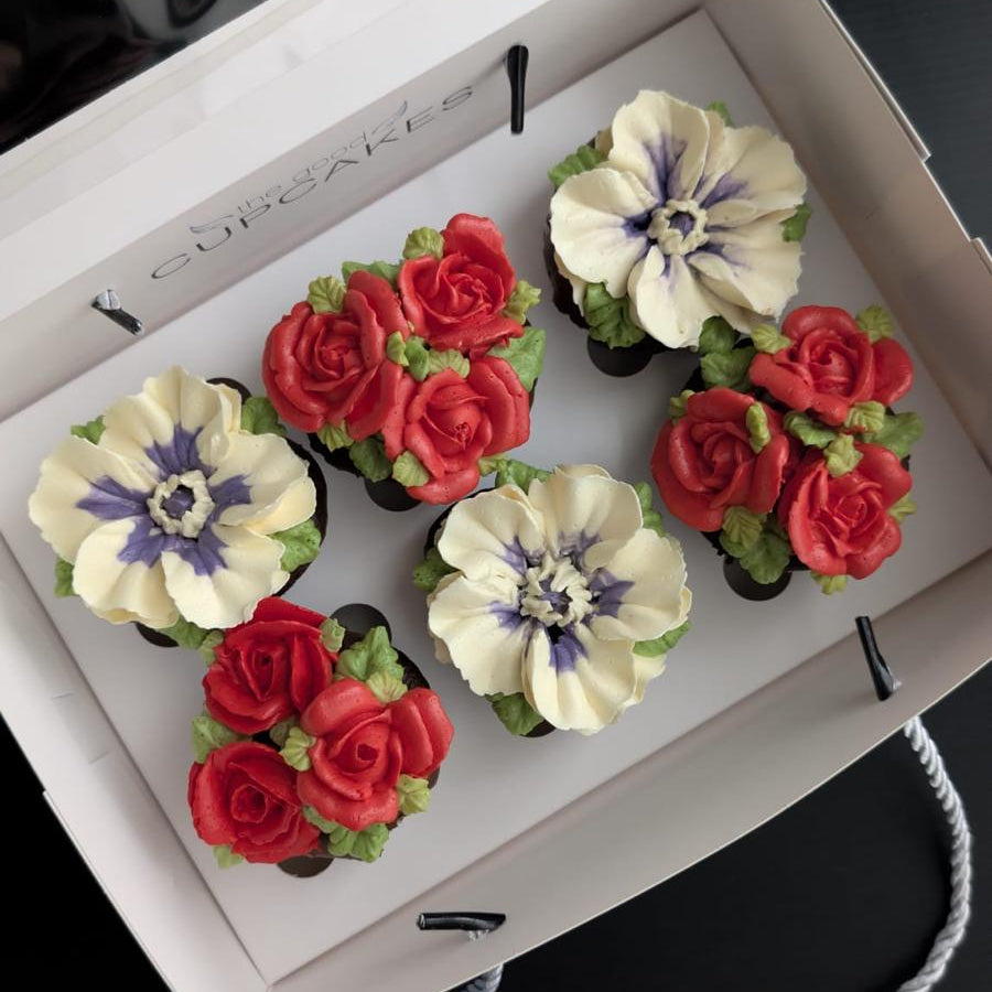 Six decorative flower cupcakes arranged inside an open white gift box on a black background. The set includes white flowers with purple centers and red rose clusters with green leaves.