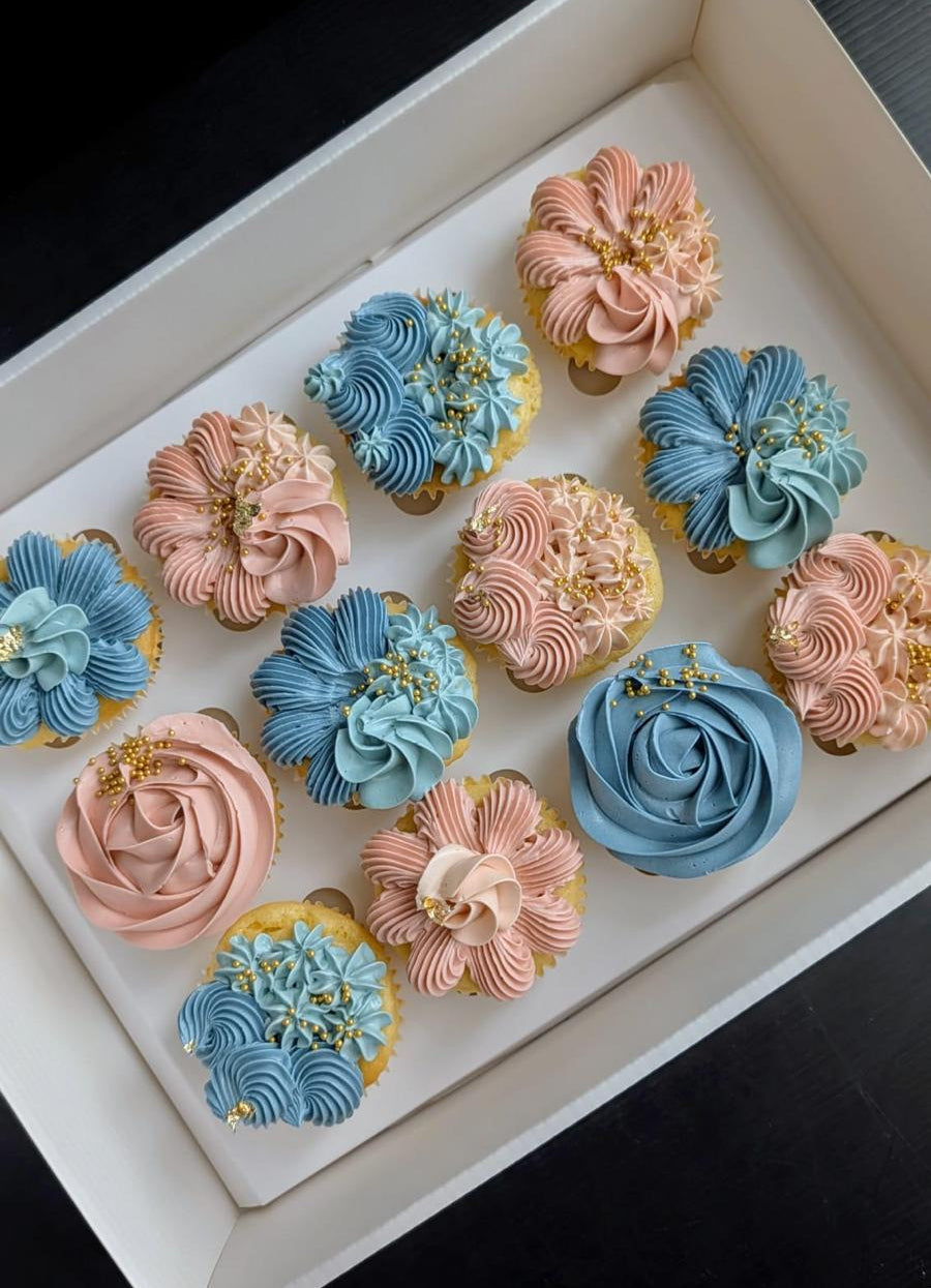 A boxed assortment of decorated gender reveal cupcakes with pink, blue, and peach frosting, arranged in a white tray on a dark background.