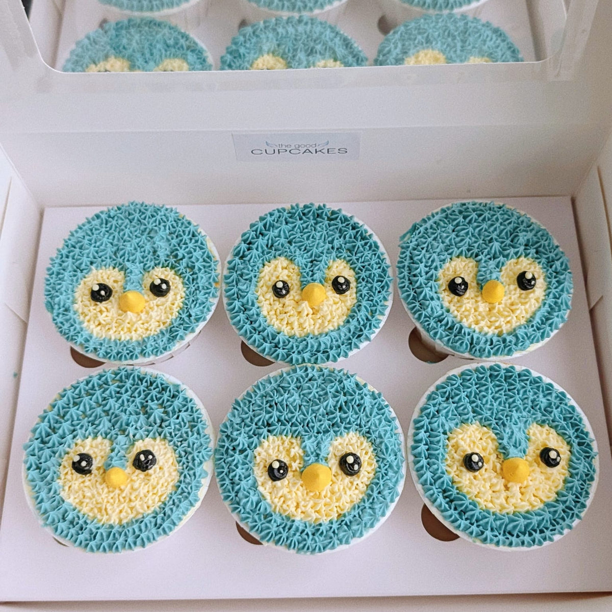 Six blue penguin cupcakes decorated with blue and yellow frosting, displayed inside a white box with a clear lid. The top of the box shows a reflection of the cupcakes.