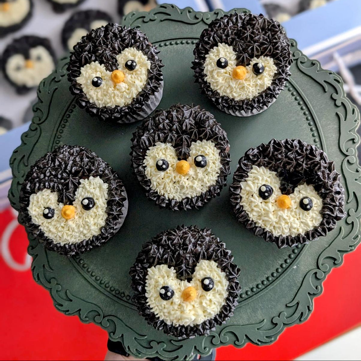 Six penguin-shaped cupcakes decorated with black frosting, white frosting, and orange frosting accents, arranged on a dark green scalloped serving platter. Two blue gift boxes containing more cupcakes are visible in the background.