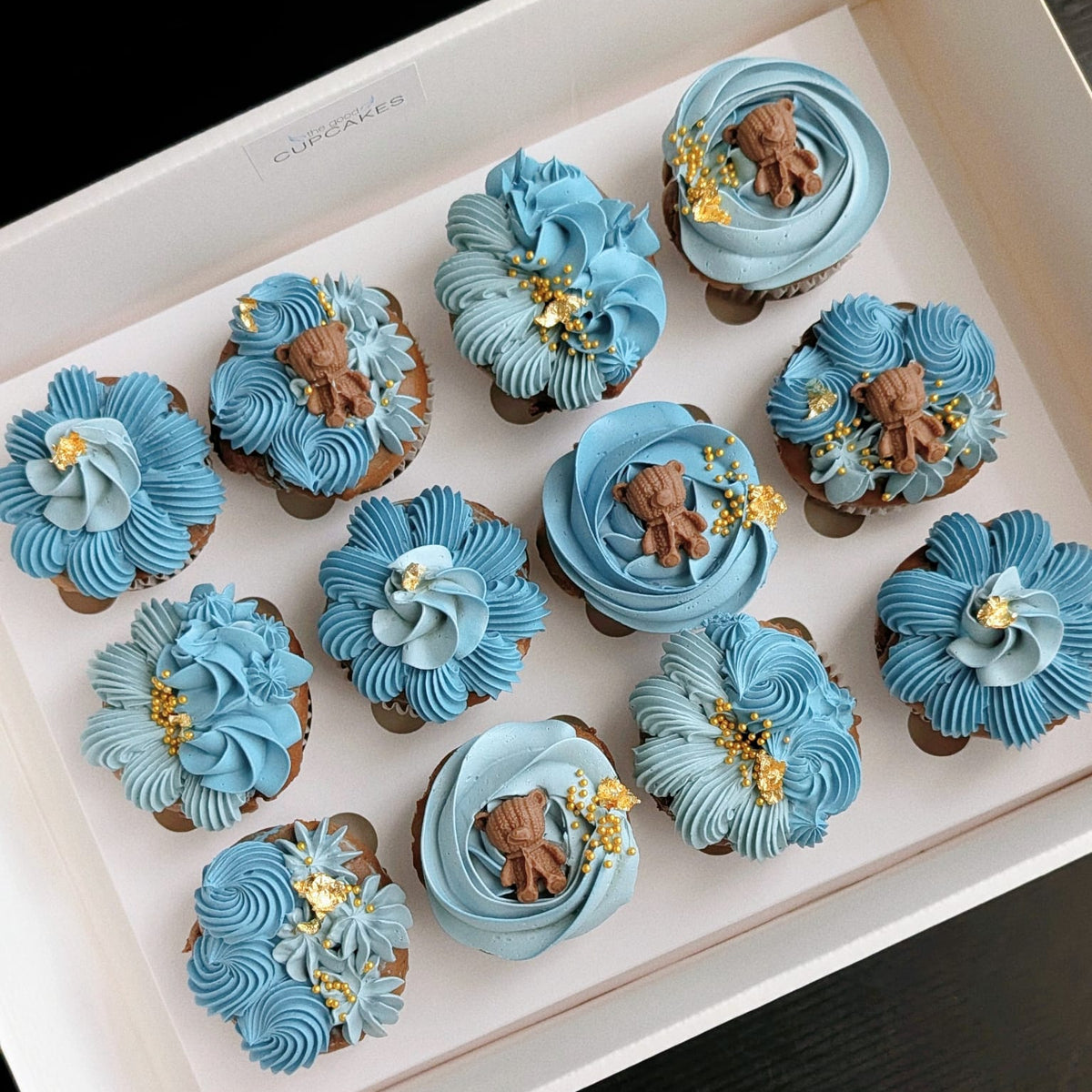 Box of 12 gender reveal cupcakes with hand-piped blue buttercream florals, teddy bear toppers and gold leaf, The Good Cupcakes Singapore