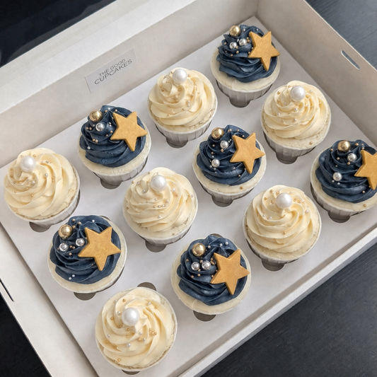 Box of 12 hand-piped cupcakes in a white gift box — six navy blue buttercream swirls topped with a gold chocolate star and gold and silver pearl sprinkles, and six ivory buttercream swirls topped with a white pearl and gold and silver dragees