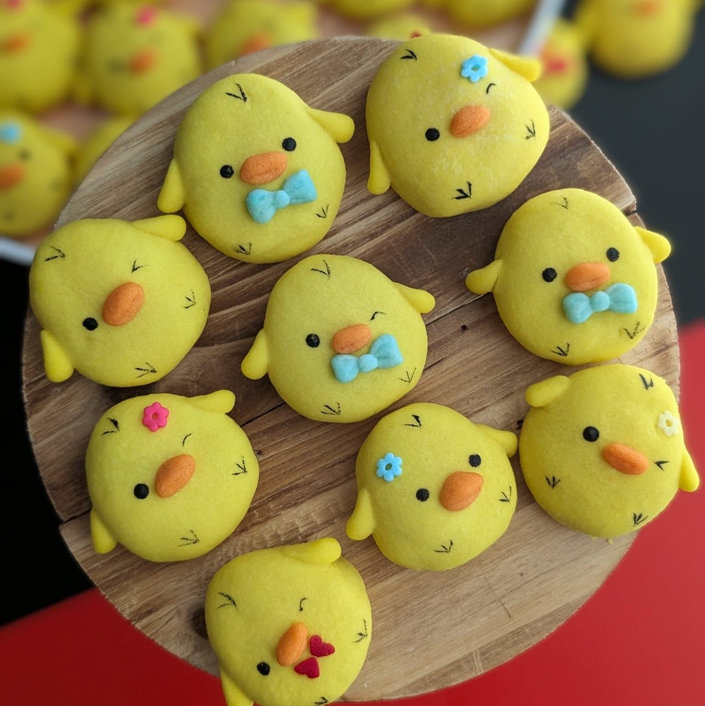 A group of yellow chick-shaped cookies decorated with colorful icing details, arranged on a round wooden board with more cookies blurred in the background.