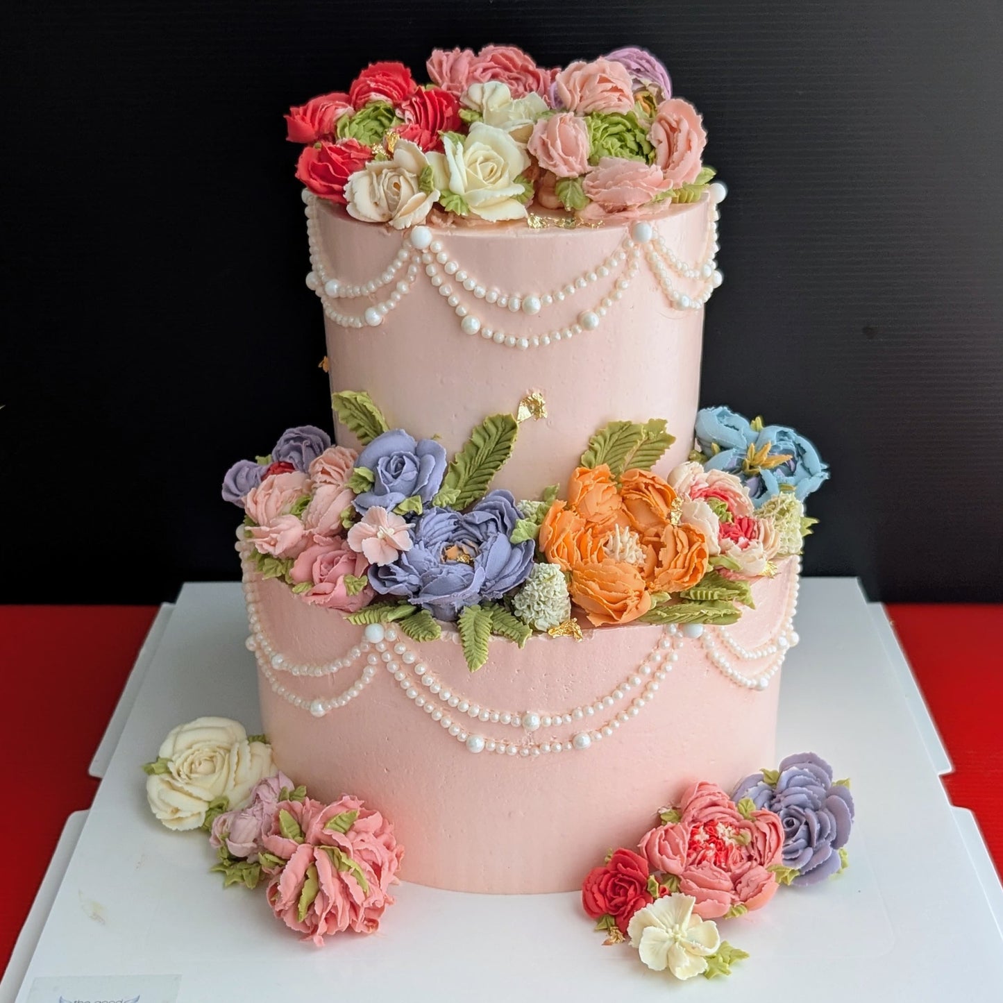 This is a two-tiered pink cake with cascading pearl draping and an abundance of hand-piped multicolour buttercream flowers (roses, peonies, ranunculus in pink, purple, blue, orange, and white).