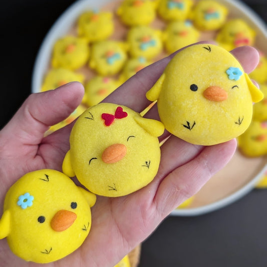 Hand-painted yellow chick-shaped shortbread cookies held in a hand, showing cute facial details with orange beaks, flower and bow decorations — low-sugar handmade cookies by The Good Cupcakes