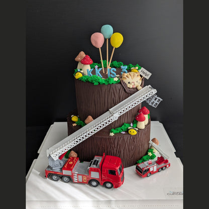 A creative two-tiered tree stump birthday cake by The Good Cupcakes Company, decorated to look like a fire station scene, featuring a red fire truck, ladder, mushrooms, balloons, and a cat topper, displayed on a white cake box against a dark background.