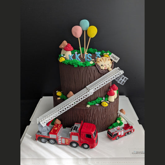 A creative two-tiered tree stump birthday cake by The Good Cupcakes Company, decorated to look like a fire station scene, featuring a red fire truck, ladder, mushrooms, balloons, and a cat topper, displayed on a white cake box against a dark background.